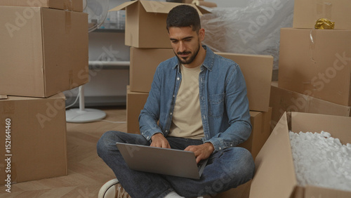 Fotografija Young man sitting on floor with laptop surrounded by moving boxes in a new home suggesting relocation or setting up in a modern living room