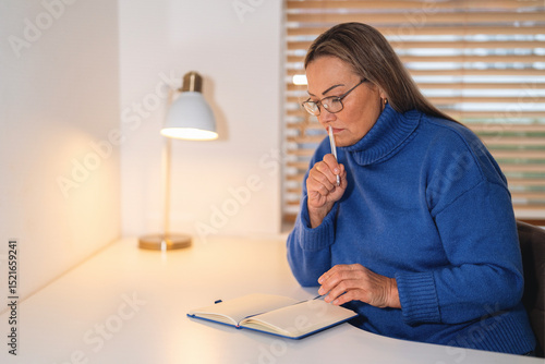 Tableau sur toile Woman in a blue sweater thoughtfully writing in a notebook at a desk in a cozy r