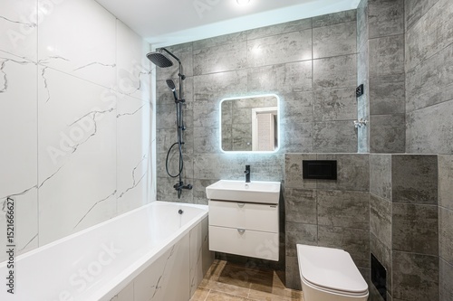 modern bathroom with white tub  sink  toilet  and a lighted mirror. Walls are tiled in white marble and gray stone