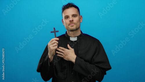 Fotografia Priest smiling holding cross on blue background, exuding calm spirituality, dressed in black robe, hands over heart in peaceful reflection, symbolizing faith and devotion