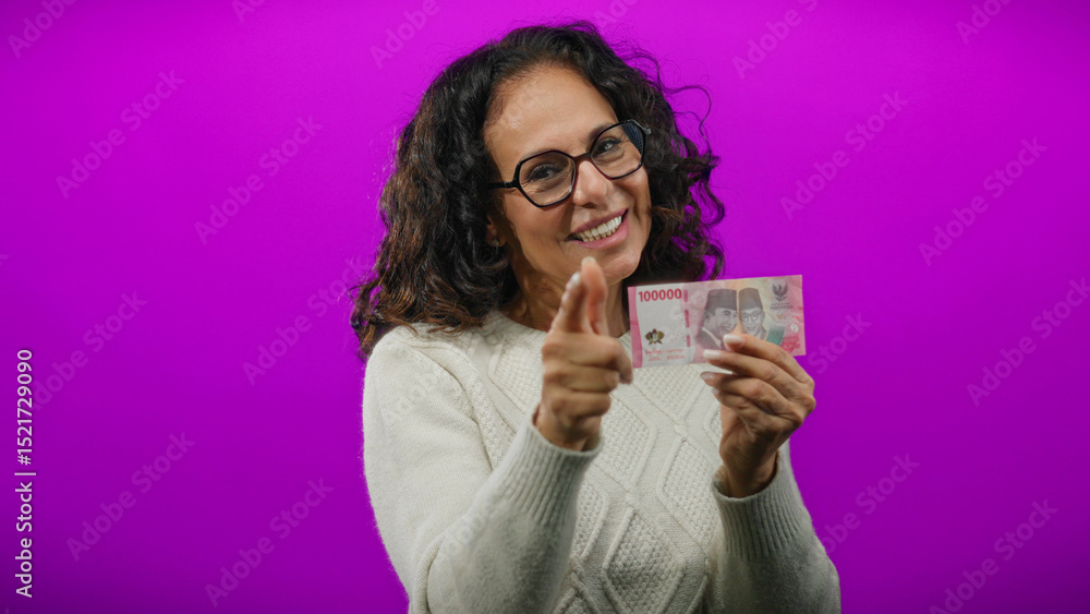 Woman smiling holding indonesian rupiah banknotes against vibrant purple background pointing at camera with confidence and positivity in a cheerful isolated setting. Woman smiling holding indonesian rupiah banknotes against vibrant purple background pointing at camera with confidence and positivity in a cheerful isolated setting.