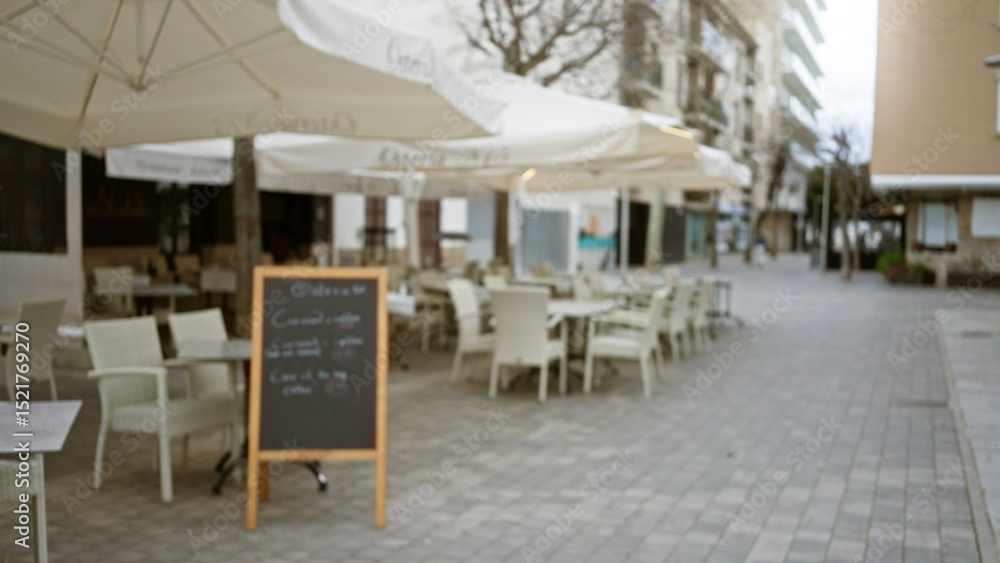 Fototapeta premium Blurred view of an outdoor cafÃ© on a cobblestone street in a mediterranean old town featuring empty chairs under large umbrellas and a blackboard menu.