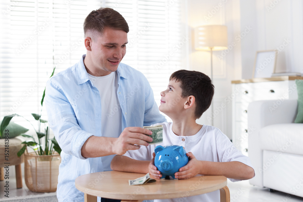 Father and son putting pocket money into piggybank at table indoors Father and son putting pocket money into piggybank at table indoors