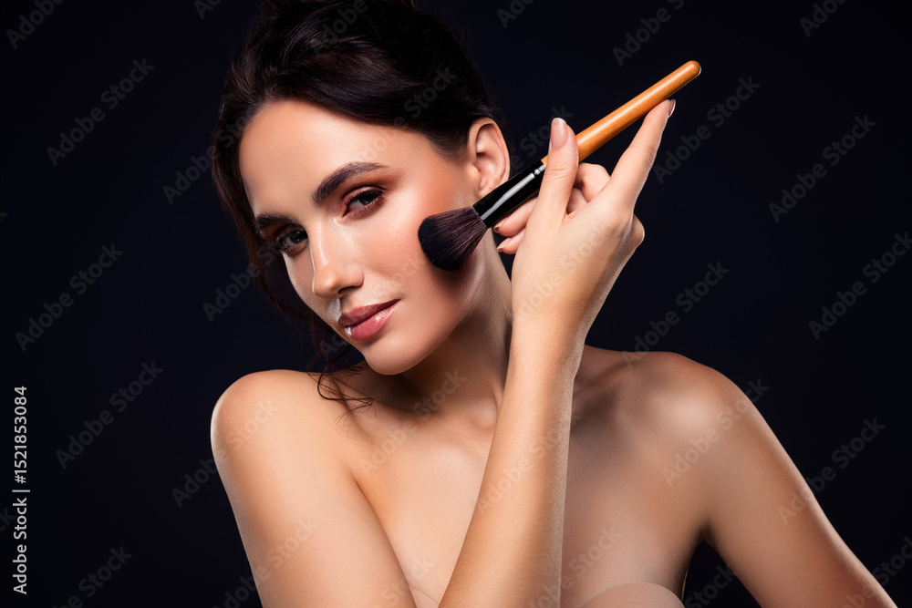 Obraz premium Young brunette woman applying professional makeup with a blush brush against a dark background