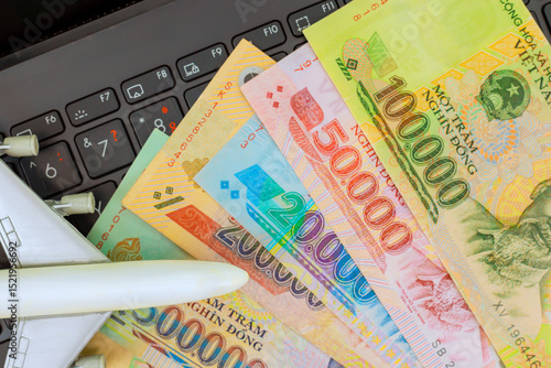 Vietnamese dong cash currency rests on laptop keyboard beside small airplane model, hinting at travel plans.