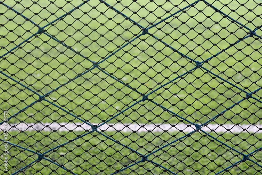 Fototapeta premium Close-up of a green sports net fence with a blurry football field in the background. Ideal for illustrating boundaries, protection, or sports facilities.