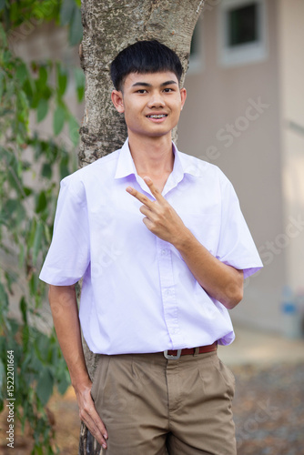 Thai boy student in official Thai school uniform (khaki or brown shorts), properly dressed, stand firmly in front of the tree with two fingers up, vertical image