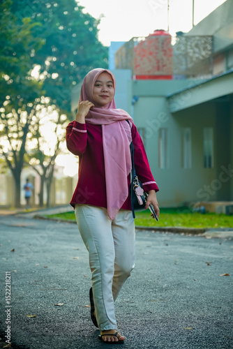 asian muslim woman facing front with slight smile walking outdoors with building background, beautiful asian muslim woman smiling and looking at camera, beautiful young asian malay woman wearing hijab