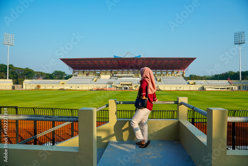 Back view of young muslim girl in hijab enjoying the view in football stadium, woman in hijab standing with her back to the camera in the empty stadium stands with a large football field spread out in