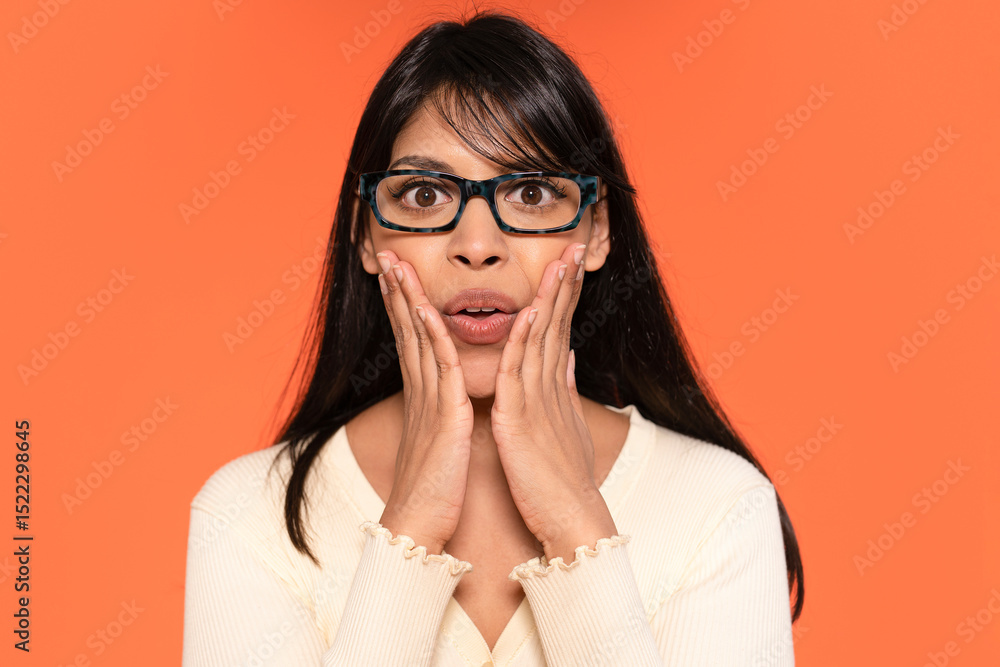 Surprised woman showing shock with hands on her face in front of an orange backdrop during a bright, sunny afternoon Surprised woman showing shock with hands on her face in front of an orange backdrop during a bright, sunny afternoon