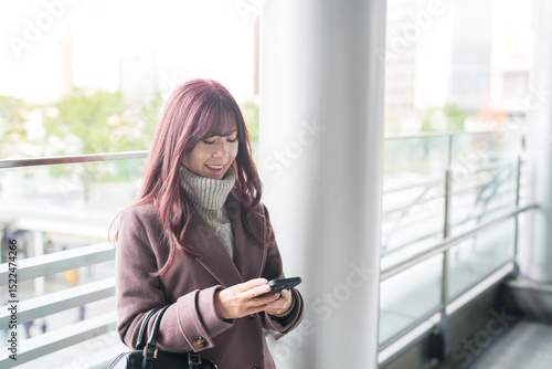 Free Download On a cold December morning, a Japanese woman in her twenties with long pink hair and a pink trench coat stands on the outdoor corridor of a large commercial building, quietly using her smartphone. - GFXLoad