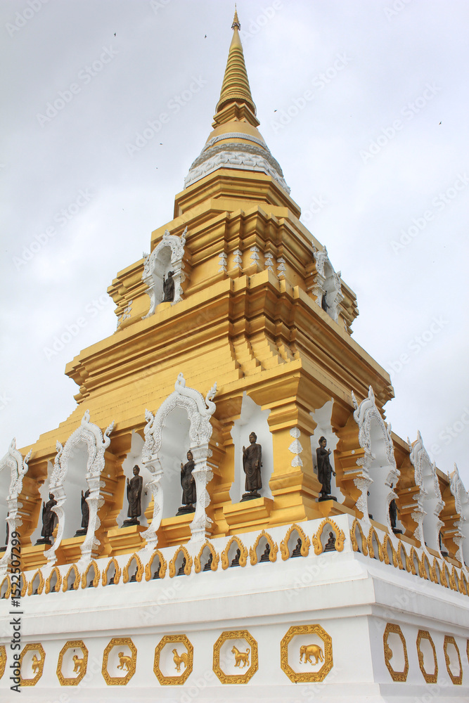 Obraz premium Golden Buddhist Pagoda in Thailand. A golden stupa decorated with Buddha statues and ornate carvings, representing Thai Buddhist architecture under cloudy sky.