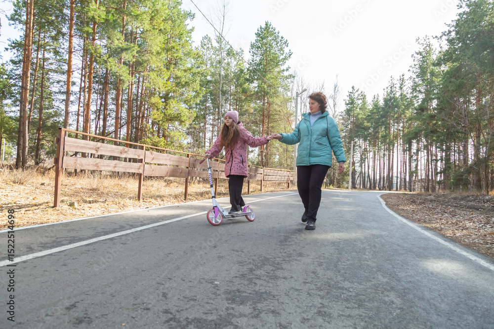 Fototapeta premium Little girl riding scooter while walking with her grandmother in the forest on a sunny day