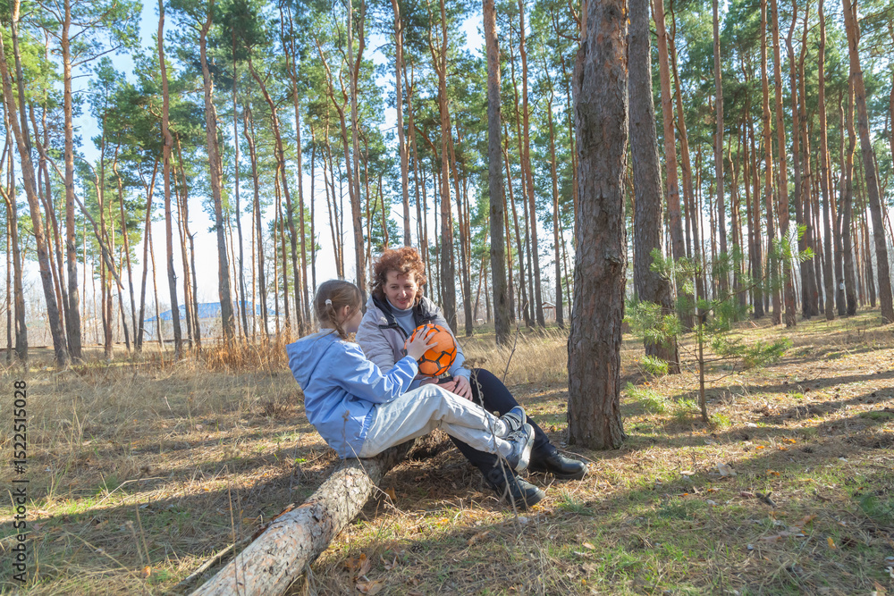 Grandmother and granddaughter talking while sitting on log in forest holding sports ball on sunny day Grandmother and granddaughter talking while sitting on log in forest holding sports ball on sunny day