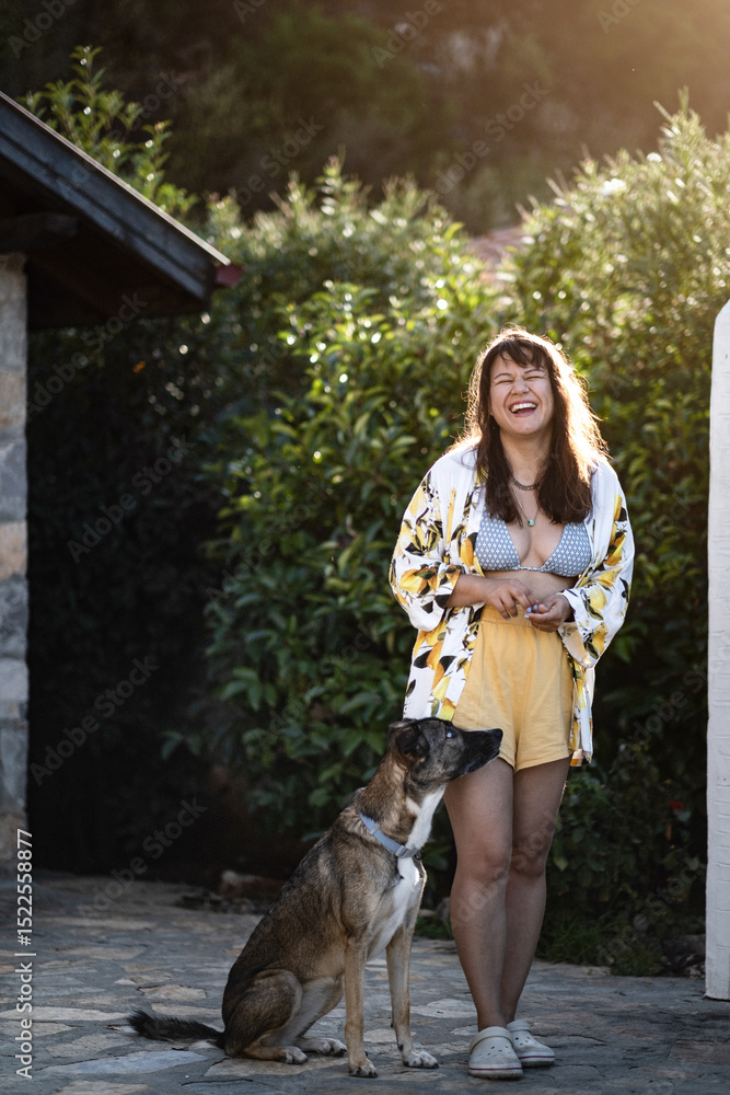 Obraz premium A woman laughs beside her calm dog in a sunny backyard with a stone house