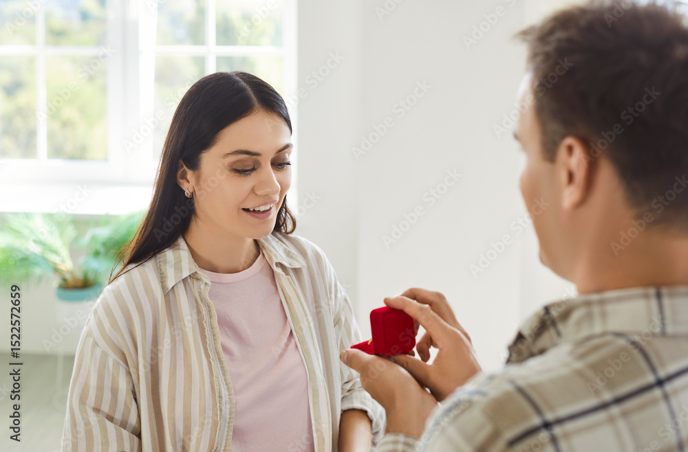 Fototapeta premium Young man holding red present box proposing marriage to happy girlfriend. Guy giving surprise gift with a engagement ring or jewelry to his cheerful girlfriend standing in living room at home.