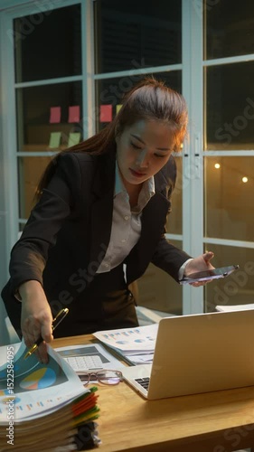 Business Asian woman in formal black suit thoughtfully reviews documents at her desk late at night, critical thinking, and commitment to meeting deadlines during late-night work hours.