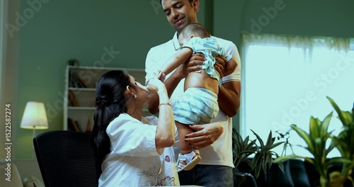 Indian mother multitasking holding infant irritated working from home on laptop with documents, phone call while father walking behind, family parenthood, young parents balancing care, stress