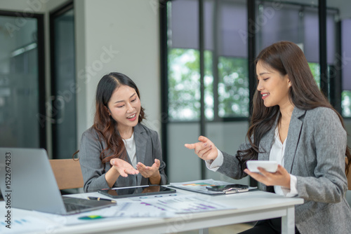 Two professional businesswomen are working on laptop computers sitting at desks in the office. Colleagues are happy and discussing the digital strategy of the Graph