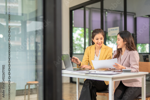 Two professional businesswomen are working on laptop computers sitting at desks in the office. Colleagues are happy and discussing the digital strategy of the Graph