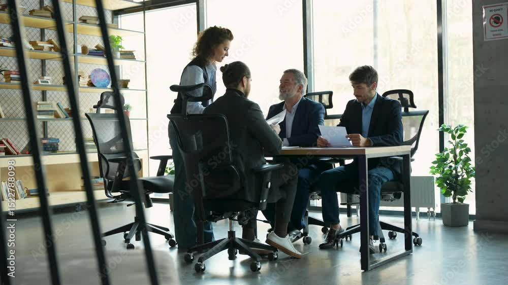 Confident female team leader boss supervisor standing at working desk holding paper documents talking to motivated interested focused teammates colleagues in modern office Confident female team leader boss supervisor standing at working desk holding paper documents talking to motivated interested focused teammates colleagues in modern office