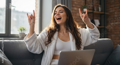 Happy young woman in bathrobe singing along to music on laptop while relaxing on couch at home