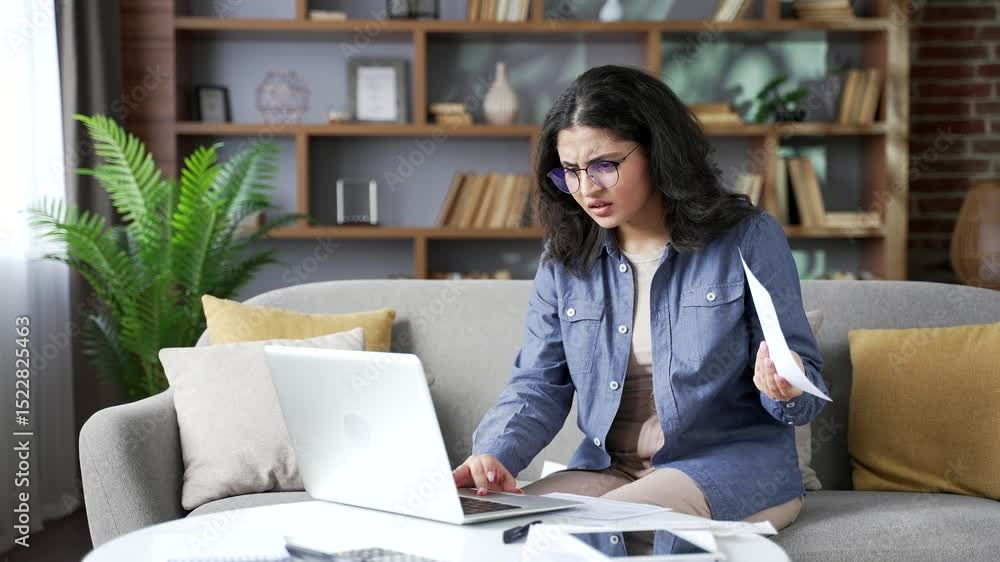 Confused young woman having difficulty doing household finances using application on laptop computer sitting on sofa in living room at home office. Puzzled shocked female looks at large utility bills Confused young woman having difficulty doing household finances using application on laptop computer sitting on sofa in living room at home office. Puzzled shocked female looks at large utility bills