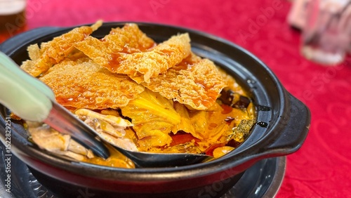Steaming Asian hotpot with crispy tofu skin, vegetables, and rich soup, served in a claypot on a traditional red dining table setup.