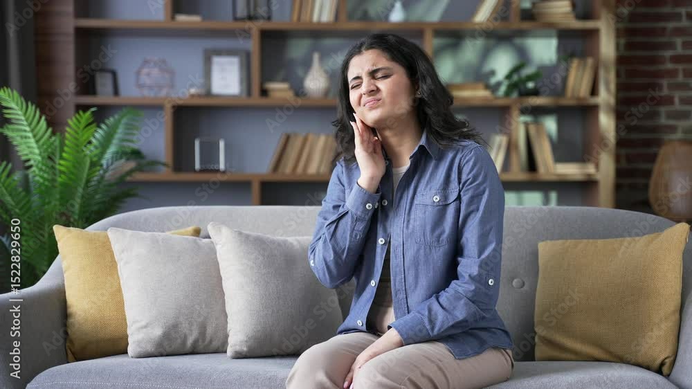 Upset young woman suffering from toothache while sitting on sofa in living room at home. The female holds her hand on her cheek, massaging the sore spot. She feels sharp pain and needs to see dentist Upset young woman suffering from toothache while sitting on sofa in living room at home. The female holds her hand on her cheek, massaging the sore spot. She feels sharp pain and needs to see dentist