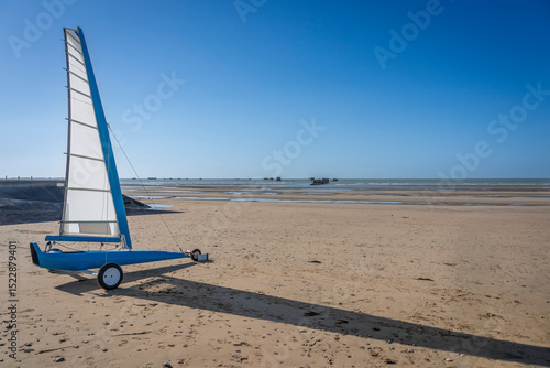 Asnelles-Sur-Mer, France - 05 16 2025:  View of a white and blue competition land yacht, a wheeled vehicle propelled by a sail along the beach between sand and sea
