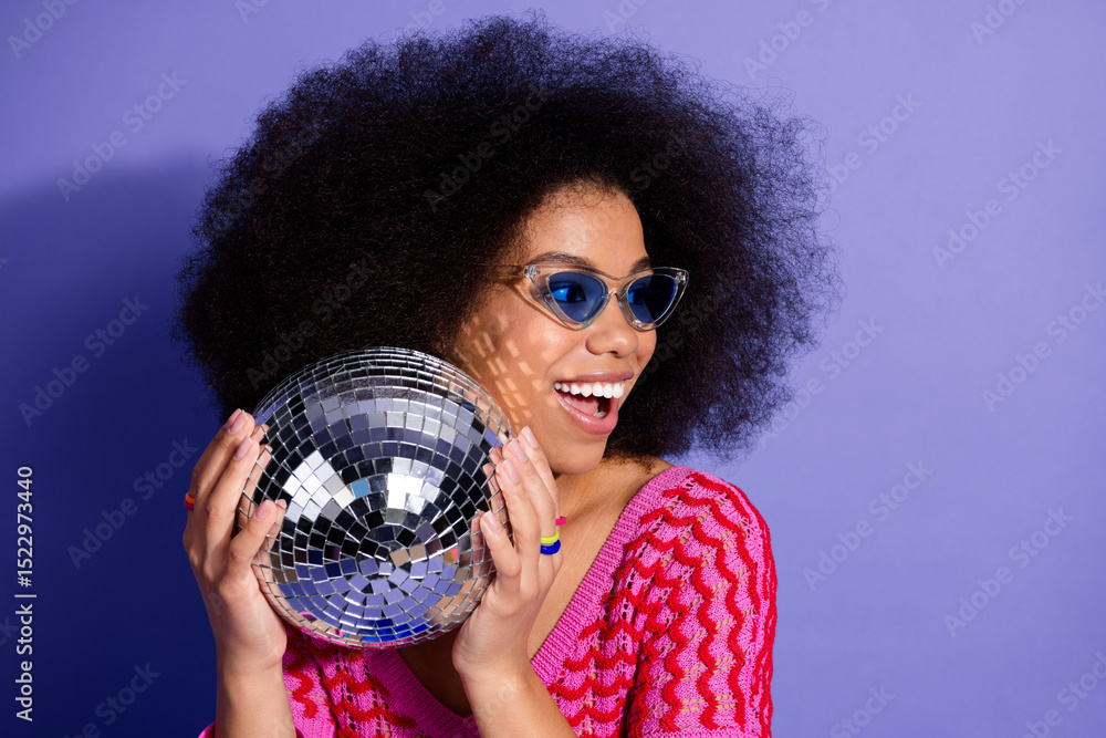 Naklejka premium Cheerful young woman in a stylish outfit holding a shiny disco ball against a vivid purple background