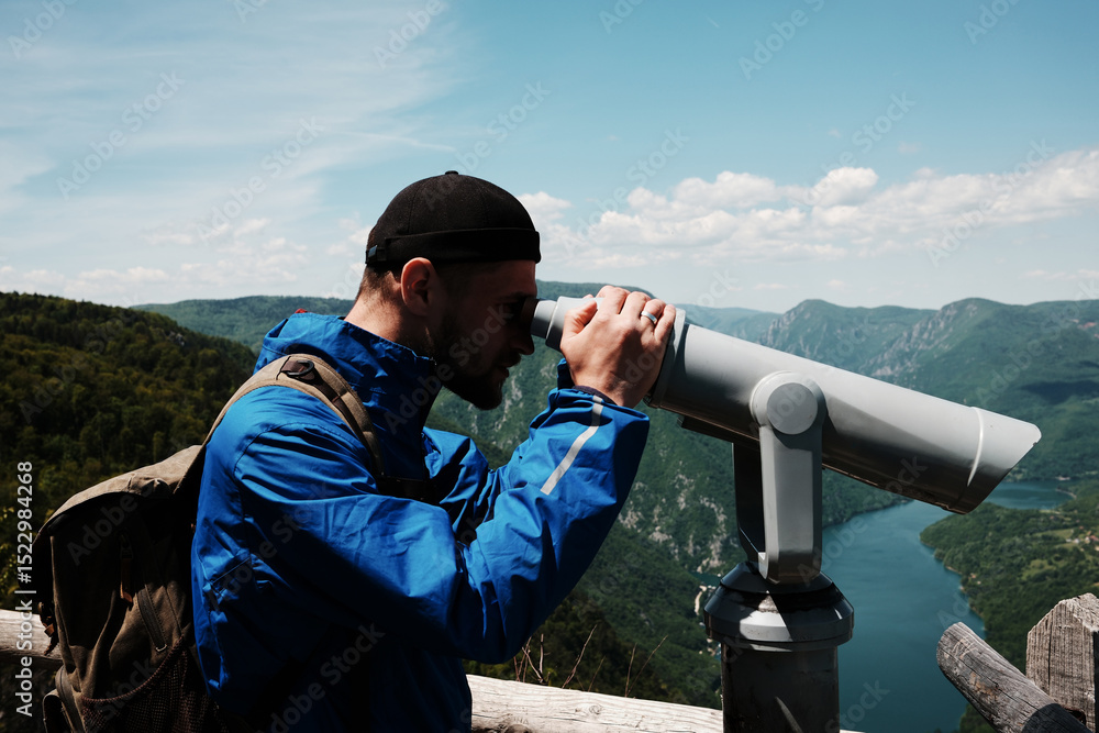 Obraz premium Man in blue jacket looking through a telescope at the lush hills and winding Drina River in Tara National Park, Serbia. Binoculars for tourists on viewpoint.