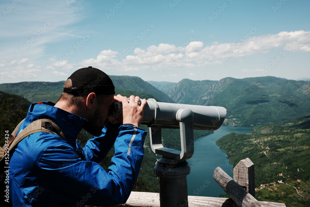 Obraz premium Man in blue jacket looking through a telescope at the lush hills and winding Drina River in Tara National Park, Serbia. Binoculars for tourists on viewpoint.