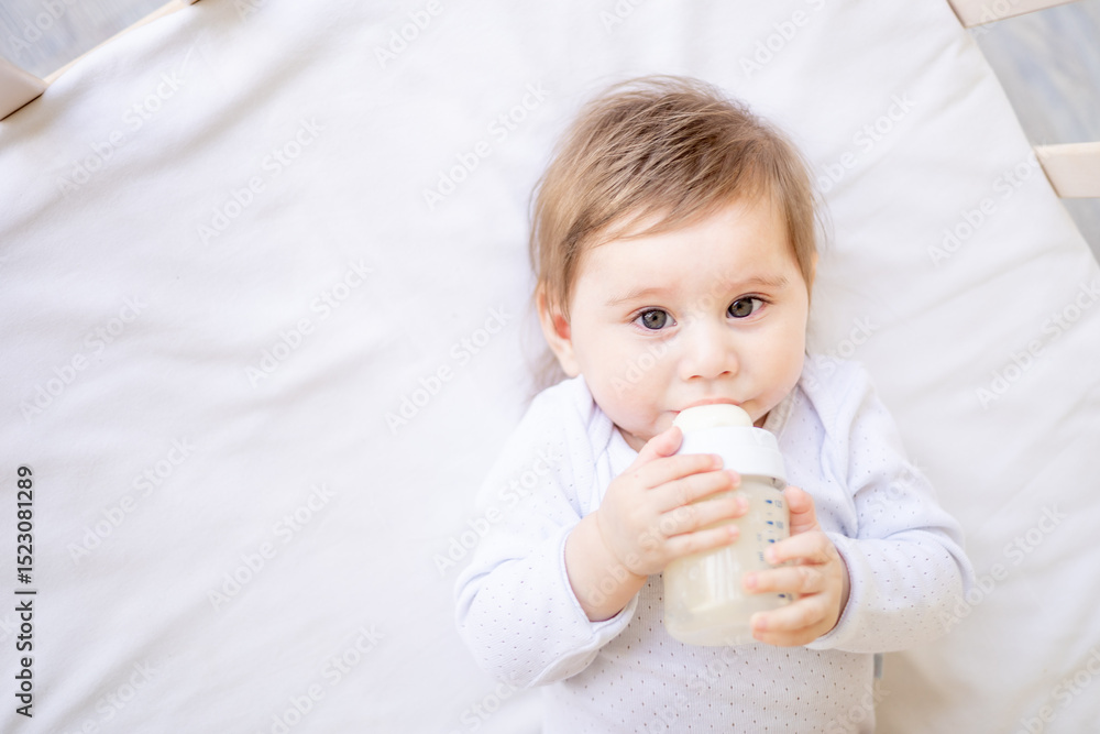 Baby newborn girl sucks a bottle of milk lying on her back in a crib in white clothes, baby food concept, close-up Baby newborn girl sucks a bottle of milk lying on her back in a crib in white clothes, baby food concept, close-up