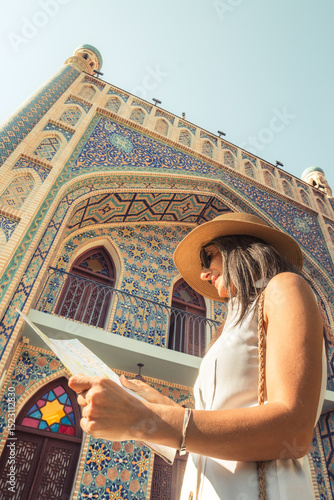 Smiling female tourist with map by colorful islamic architecture Persian-style mosque building. Travel sightseeing planning, cultural discovery, solo journey, heritage adventure