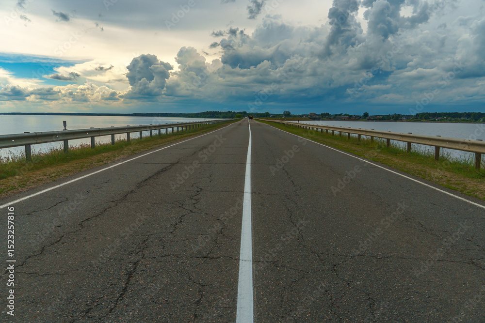 Fototapeta premium An empty asphalt road runs through the river. Thick gray clouds over the highway along the lake.