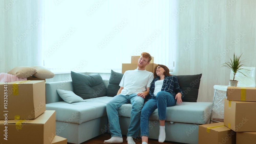 Caucasian joyful boyfriend and girlfriend moving in together in new house. Tired happy young married couple sitting down on sofa in room with many boxes on moving day. Home concept Caucasian joyful boyfriend and girlfriend moving in together in new house. Tired happy young married couple sitting down on sofa in room with many boxes on moving day. Home concept