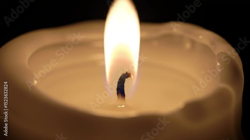 Close up of a white candle with a bright flame burning against a dark black background glow softly