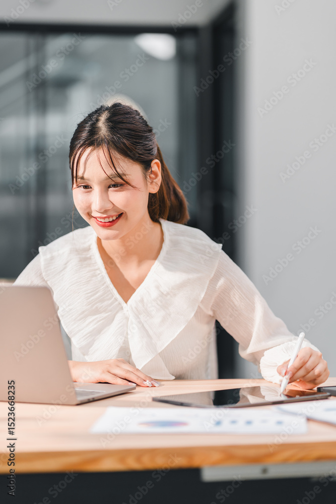 Young Asian woman in white blouse is smiling while working on laptop and using digital tablet with stylus pen at wooden desk in bright modern office environment Young Asian woman in white blouse is smiling while working on laptop and using digital tablet with stylus pen at wooden desk in bright modern office environment