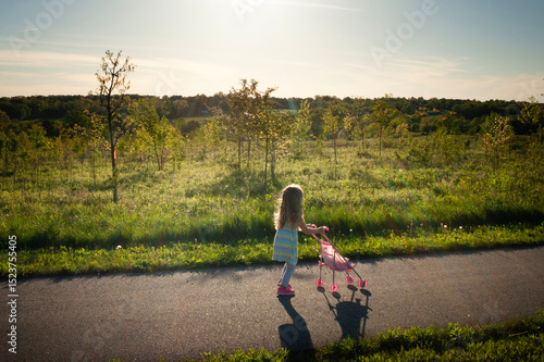 Side view young girl pushing pink baby doll stroller sunny green