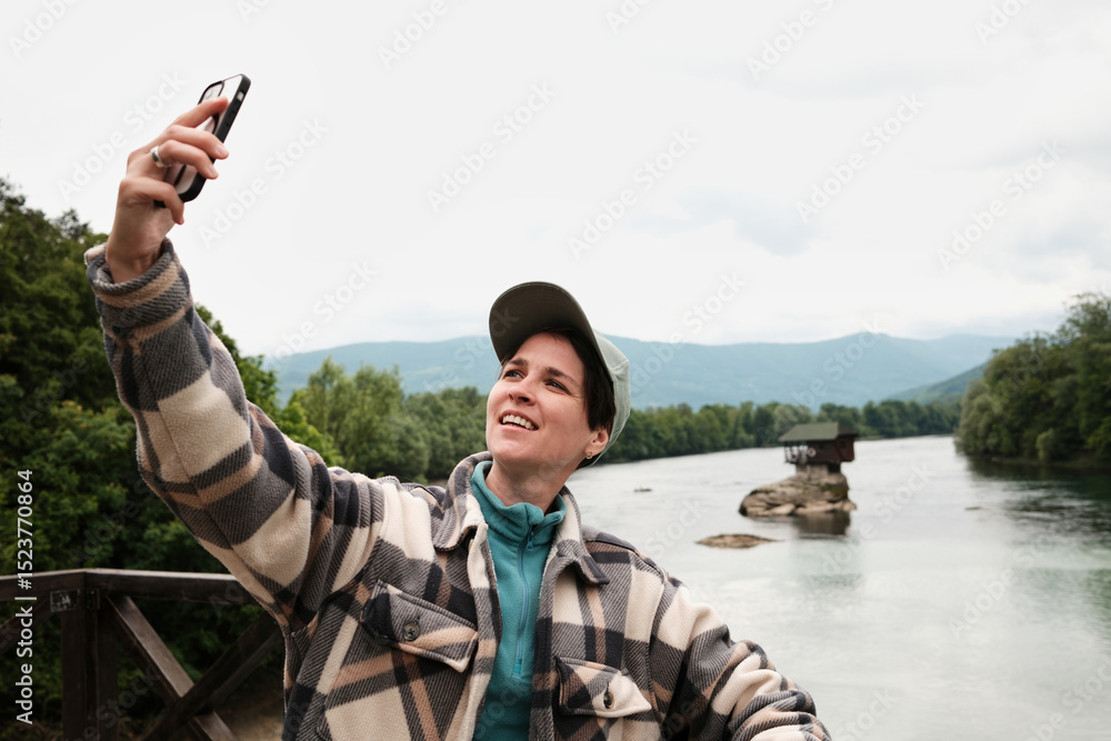 Obraz premium Woman taking a cheerful selfie by the river in front of the famous Drina house on the rock in Serbia. Female travel blogger.
