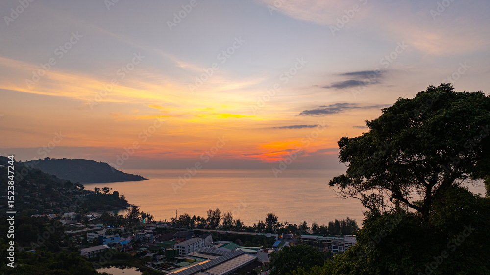 Fototapeta premium A stunning aerial view of the Andaman Sea at sunset, overlooking a coastal town in Phuket, Thailand. The golden hues of the sky reflect beautifully on the calm ocean waters, creating a tranquil