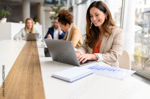 Smiling businesswoman working on project over laptop with colleagues during collaborative meeting in office