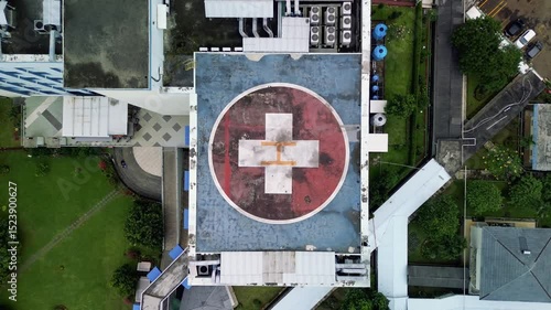 This footage presents an aerial, top-down view of a hospital helipad marked with a red cross. The helipad is surrounded by buildings, green lawns, trees, and a parking area.