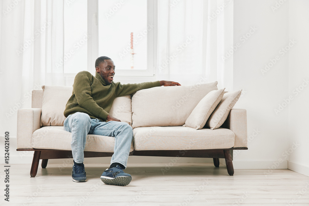 Comfortable young Black man relaxing on a beige sofa in a bright and modern living room, wearing casual clothing, with a calm and satisfied expression Comfortable young Black man relaxing on a beige sofa in a bright and modern living room, wearing casual clothing, with a calm and satisfied expression