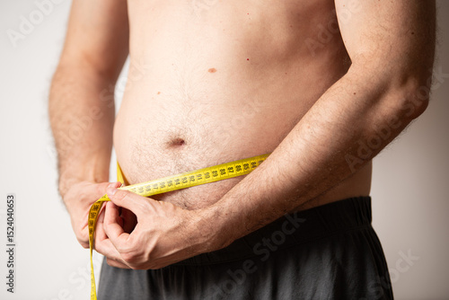 A man stands at home measuring his waist with a tape measure, focusing on his health and fitness goals. He appears determined to track his body changes