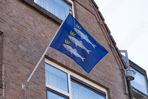 Quadro em tela A blue flag with three white fish and crowns, representing Scheveningen, flies against a brick building, symbolizing local heritage and identity