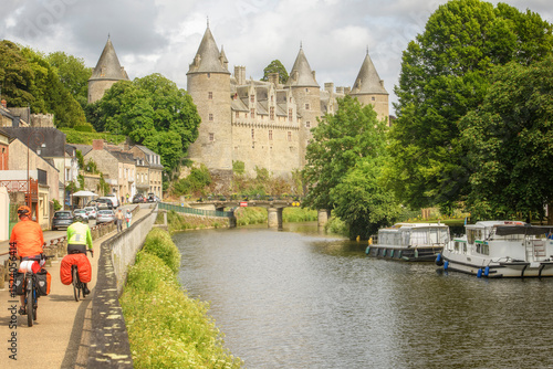 Couple en vélo le long d'une riviere avec des peniches et un chateau en arriere plan , velotourisme le long de la riviere Oust sur le canal de Nantes à Brest, ville de  Josselin en région Bretagne.
