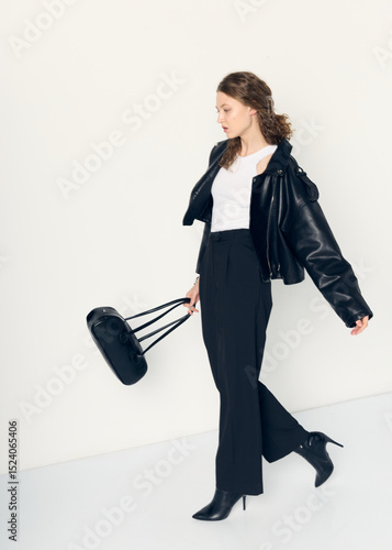 curly-haired elegant woman dressed in office style, in black pants, with a leather jacket and a bag in her hand. Posing relaxed on a white background
