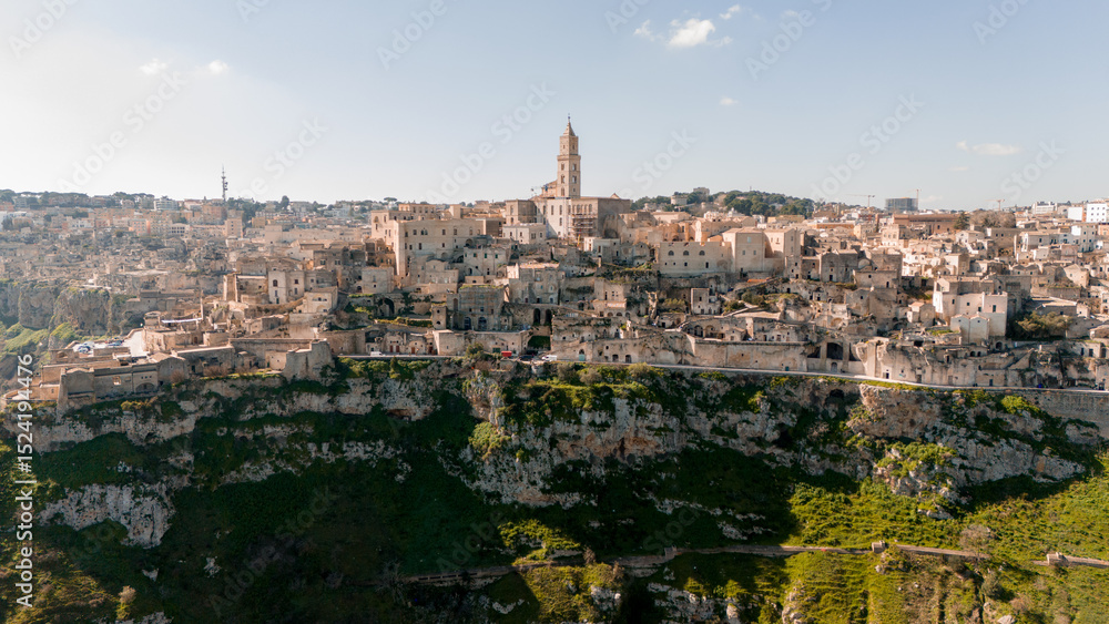 Fototapeta premium Aerial view of Matera, Italy, with its stone-built houses and cathedral tower rising above the deep ravine. A bright, clear day highlights the timeless beauty of this ancient city
